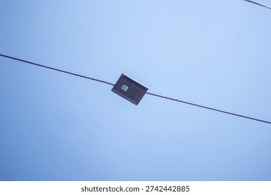 A small, dark-colored, square-shaped object with a circular detail is suspended from a thin, dark wire that stretches across a clear blue sky with a subtle gradient effect