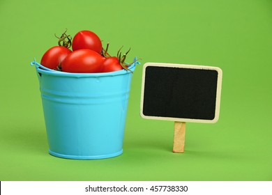 Small Disproportional Blue Bucket Of Red Cherry Tomatoes And Black Chalkboard Sign Over Green Background, Close Up