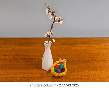 Small Easter decoration in form of tiny textile Easter basket with Easter eggs and live blossom apricot branch in a small white ceramic vase on a wooden table on a gray background