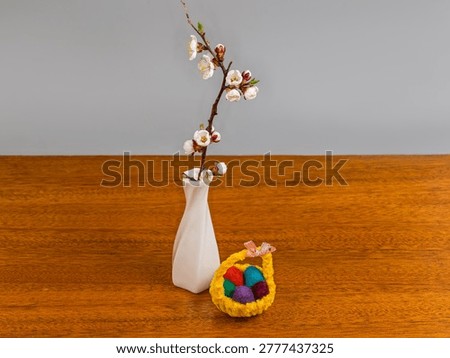 Small Easter decoration in form of tiny textile Easter basket with Easter eggs and live blossom apricot branch in a small white ceramic vase on a wooden table on a gray background
