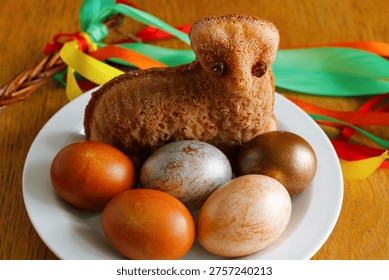 Small Easter lamb cake with painted eggs on white plate and traditional Czech pomlazka with colorful ribbons in background, festive spring holiday food still life