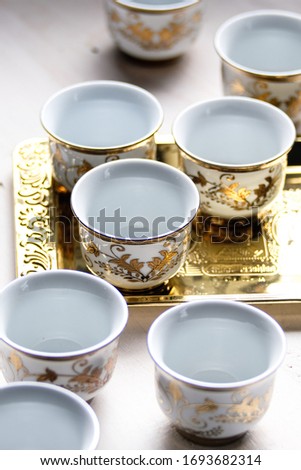 Small, empty ceramic glasses adorned on the outside with gold-colored Arab embos. Placed in a golden tray. Arranged randomly in diagonal lines in bright light.