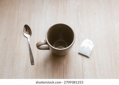 A small empty cup sits on a light wooden table next to a used tea bag and a silver spoon, indicating a moment of tea enjoyment just passed.