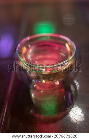 Small empty cup with transparent double glass bottom on wooden surface, object shot, selective soft focus. Glassware for hot beverages over blurred background, colorful neon light. Clean teacup