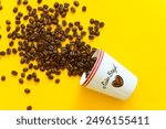 Small espresso cup on the side, with whole coffee beans rolling out of it, on a yellow background. Delicious, fresh beans in a white cup. Copy space around.