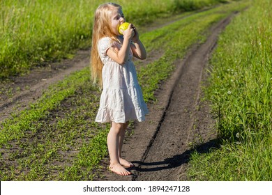 A Small Fair Girl With Long Loose Hair Stands On The Village Road Between The Fields With An Apple In Her Hands And Winces From The Bright Summer Sun Full Length In A Light Light Dress