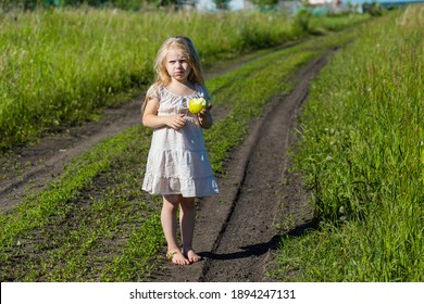 A Small Fair Girl With Long Loose Hair Stands On The Village Road Between The Fields With An Apple In Her Hands And Winces From The Bright Summer Sun Full Length In A Light Light Dress