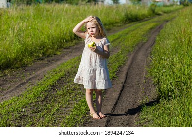 A Small Fair Girl With Long Loose Hair Stands On The Village Road Between The Fields With An Apple In Her Hands And Winces From The Bright Summer Sun Full Length In A Light Light Dress