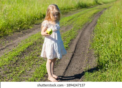 A Small Fair Girl With Long Loose Hair Stands On The Village Road Between The Fields With An Apple In Her Hands And Winces From The Bright Summer Sun Full Length In A Light Light Dress