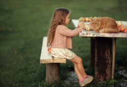 A Small, Fair-haired Girl Is Sitting On A Bench With Her Hands On The Table, Holding An Apple In Her Hands, And A Red Cat Is Next To Her. .Image With Selective Focus. The Image Is Tinted. The Image Co
