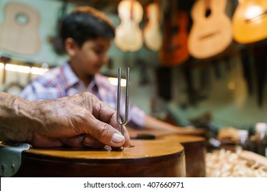 Small Family Business And Traditions: Old Grandpa With Grandson In Lute Maker Shop. The Senior Artisan Teaches How To Tune A Classic Guitar To The Boy. Closeup Of Hand Holding A Diapason 