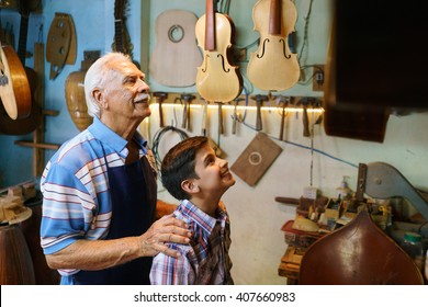 Small Family Business And Traditions: Old Grandpa With Grandson In Lute Maker Shop. The Senior Artisan Hugs The Boy And Shows Him His Handmade Guitar And Music Instruments.