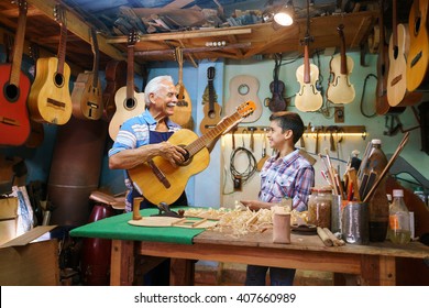 Small Family Business And Traditions: Old Grandpa With Grandson In Lute Maker Shop. The Senior Artisan Gives Teaches How To Play Classic Guitar To The Boy, Who Looks Carefully At The Instrument