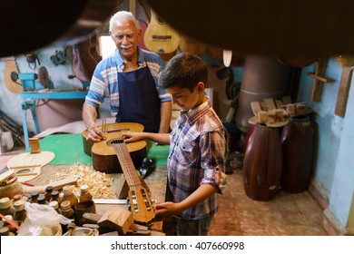 Small Family Business And Traditions: Old Grandpa With Grandson In Lute Maker Shop. The Senior Artisan Teaches How To Tune A Classic Guitar To The Boy, Holding A Diapason.