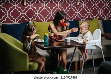 Small Family Eating In A Cafe. Mom And Her Two Children. Sitting On A Sofa, Feeding Infant Child In A Baby Chair With The Spoon.