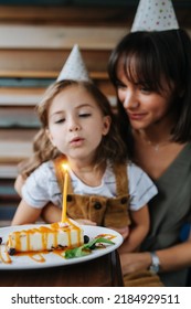 Small Family Holiday. Mom Celebrating Her Daughter's Birthday With Her Both Wearing Party Cones, Blowing A Candle. In A Cafe.