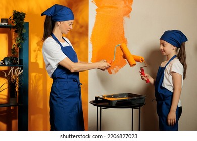 Small Family Painting Orange Walls At Home, Using Color Paint And Roller Paintbrush To Work On Apartment Renovation. Woman With Little Child Redecorating House Interior With Tools.