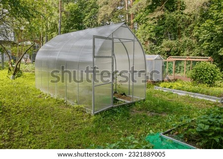 Small farm greenhouse with open door for growing plants on a hothouses for cultivation of tomatoes, cucumbers and peppers with strawberry bed patch background in yard.

Farming and gardening concept.
