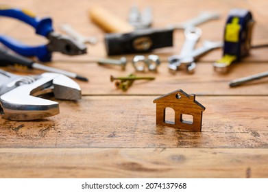 Small Figurine Of A House On The Background Of Construction Tools On A Wooden Table. House Construction And Renovation Concept. Selective Focus On A Small House.