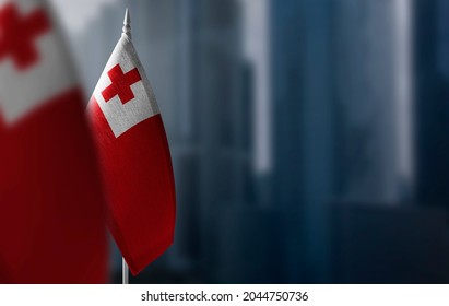 Small Flags Of Tonga On A Blurry Background Of The City.