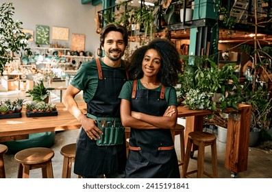 Small flower business. Front view of caucasian man and african american woman in aprons smiling at camera white standing at own shop. Happy couple selling fresh green plants for home decor. - Powered by Shutterstock