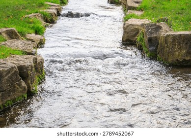 Small flowing stream bordered by natural stones and green grass in a landscaped park with moving water and peaceful outdoor scenery