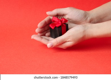 A Small Gift Box With A Red Ribbon On A Bright Red Background. Women's Hands Hold A Gift. Christmas, February 14, Birthday. Focus On The Gift.