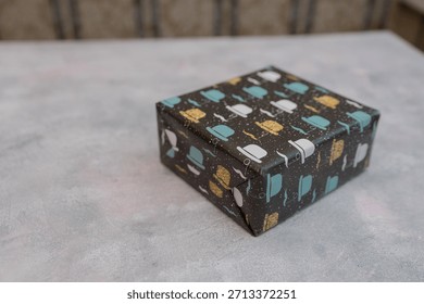 A small gift box wrapped in patterned paper featuring colorful teapots and cups. The box is placed on a light gray surface.
