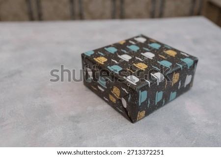 A small gift box wrapped in patterned paper featuring colorful teapots and cups. The box is placed on a light gray surface.