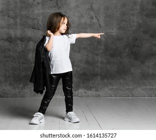 Small Girl In Black And White Rock Star Style Casual Clothing And White Sneakers Standing And Pointing Aside With Finger Over Grey Concrete Background In Studio. Stylish Children Clothing Concept