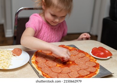 Small Girl Cooking Food. Little Child Making Pizza, Sitting At Table In Kitchen At Home, Putting Pepperoni On Dough.