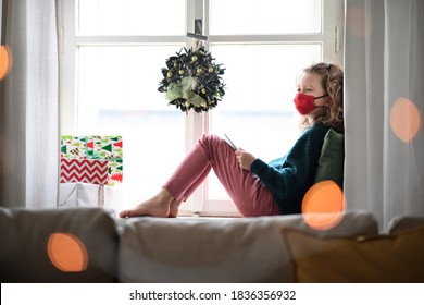 Small girl with face mask sitting indoors at home at Christmas, using tablet. Coronavirus concept.