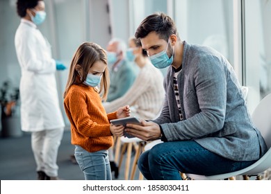 Small girl and her father with face masks using touchpad while siting in waiting room at medical clinic. - Powered by Shutterstock