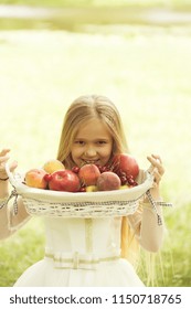 Small Girl Kid With Long Blonde Hair And Pretty Smiling Happy Face In Prom Princess White Dress Standing Sunny Day Outdoor Near Water With Fruit Basket Of Red Apples Peach And Cherry.