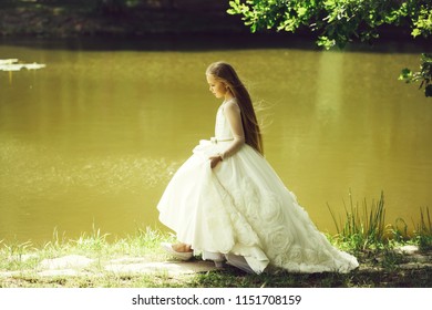 Small Girl Kid With Long Blonde Hair And Pretty Smiling Happy Face In Prom Princess White Dress Standing Sunny Day Outdoor Near Water