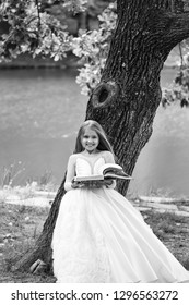 Small Girl Kid With Long Blonde Hair And Pretty Smiling Happy Face In Prom Princess White Dress Standing Sunny Day Outdoor Near Water With Book