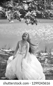 Small Girl Kid With Long Blonde Hair And Pretty Smiling Happy Face In Prom Princess White Dress Standing Sunny Day Outdoor Near Water