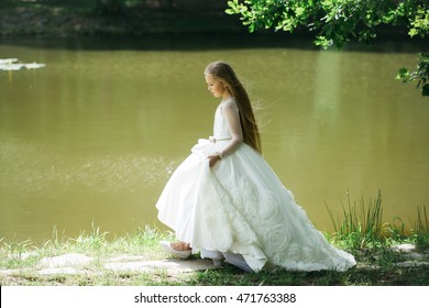 Small Girl Kid With Long Blonde Hair And Pretty Smiling Happy Face In Prom Princess White Dress Standing Sunny Day Outdoor Near Water