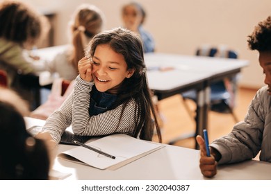Small girl laughs happily in a primary school class, she is sitting at a table with her her classmates. Elementary school students enjoying an art and creativity class in a child development centre.