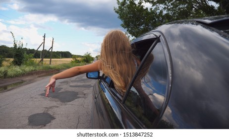 Small Girl Leaning Out Of Car Window And Waving Her Hand In Wind While Riding Through Country Road. Little Child Looking Out Of Open Window Moving Auto And Her Arm Playing With Breeze. Rear View