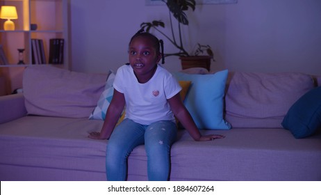 Small Girl On Couch Watching Tv At Night In Living Room. Cute Afro-american Kid Sitting Down On Sofa And Watching Cartoons Late In Evening. Childhood Ad Entertainment Concept