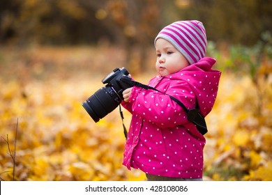 A small girl is photographer, kid holds a camera and takes photo of autumn landscape, child stands in the autumn park and girl is dressed in warm hat and jacket.