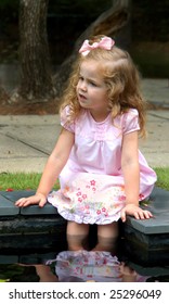 Small Girl Sits On Edge Of Lily Pad Pool And Wets Her Feet.  She Is Frowning And Confused.  Pink Dress And Hairbow.