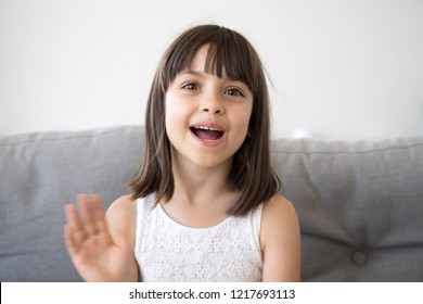 Small girl sitting on couch alone at home. Head shot portrait little adorable daughter waving hand looking at camera smile saying hello or goodbye. First acquaintance and online communication concept
