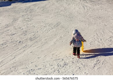 Small Girl In Winter Clothing Pushes A Wooden Sledge Through To The Top Of A Snow Covered Hill. Seen From A Low Perspective.