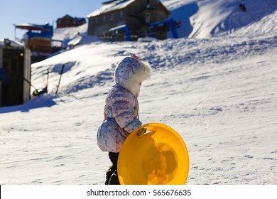 Small Girl In Winter Clothing Pushes A Wooden Sledge Through To The Top Of A Snow Covered Hill. Seen From A Low Perspective.