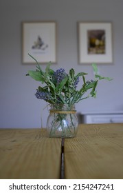 A Small Glass Pot Of Purple And Blue Flowers And Green Leaves, On A Wooden Table. Isolated On Its Own. Leading Line Down The Middle. Picture Frames With Artwork In The Background. No People. Vertical.