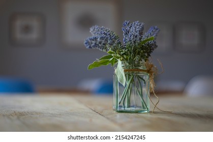 A Small Glass Pot Of Purple And Blue Flowers And Green Leaves, On A Wooden Table. Isolated On Its Own. Scandi Chic Minimalism Interior Design. Picture Frames With Artwork In The Background. No People.