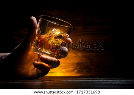 a small glass with whiskey and ice in a male hand on a background of a wooden wall with place for text. a ray of light shines on a glass
