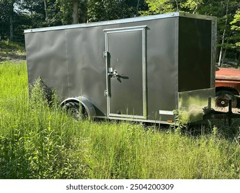 Small gray, enclosed trailer, parked in high grass next to an old brown SUV. - Powered by Shutterstock